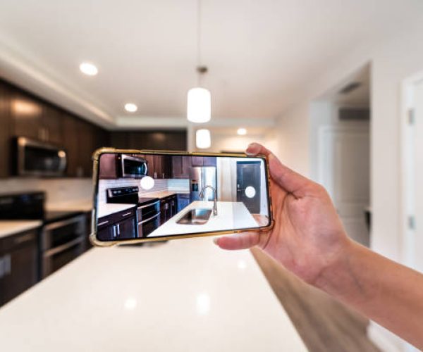 istockphoto-1404958581-612×612 Hand photographing house apartment kitchen island room for sale or rent tour with phone smartphone closeup point of view in modern luxury condo home with blurry bokeh background